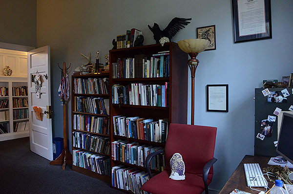 Overview of office from corner with windows, facing desk, wall of bookshelves, and the door