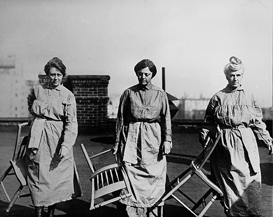 Outdoor photograph of three National Woman’s Party members in prison dress carrying wooden chairs