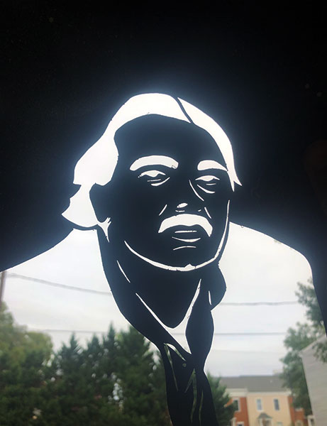 A stencil depicting poet Larry Levis held up with sky and houses showing through the stencil.