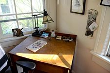 Emerson’s desk angled in a corner by a window; lamp, paperweight, open face-down Dickinson volume among items on desk.