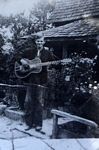 B&W unframed image of a man playing guitar in front of a wood-shingled house.