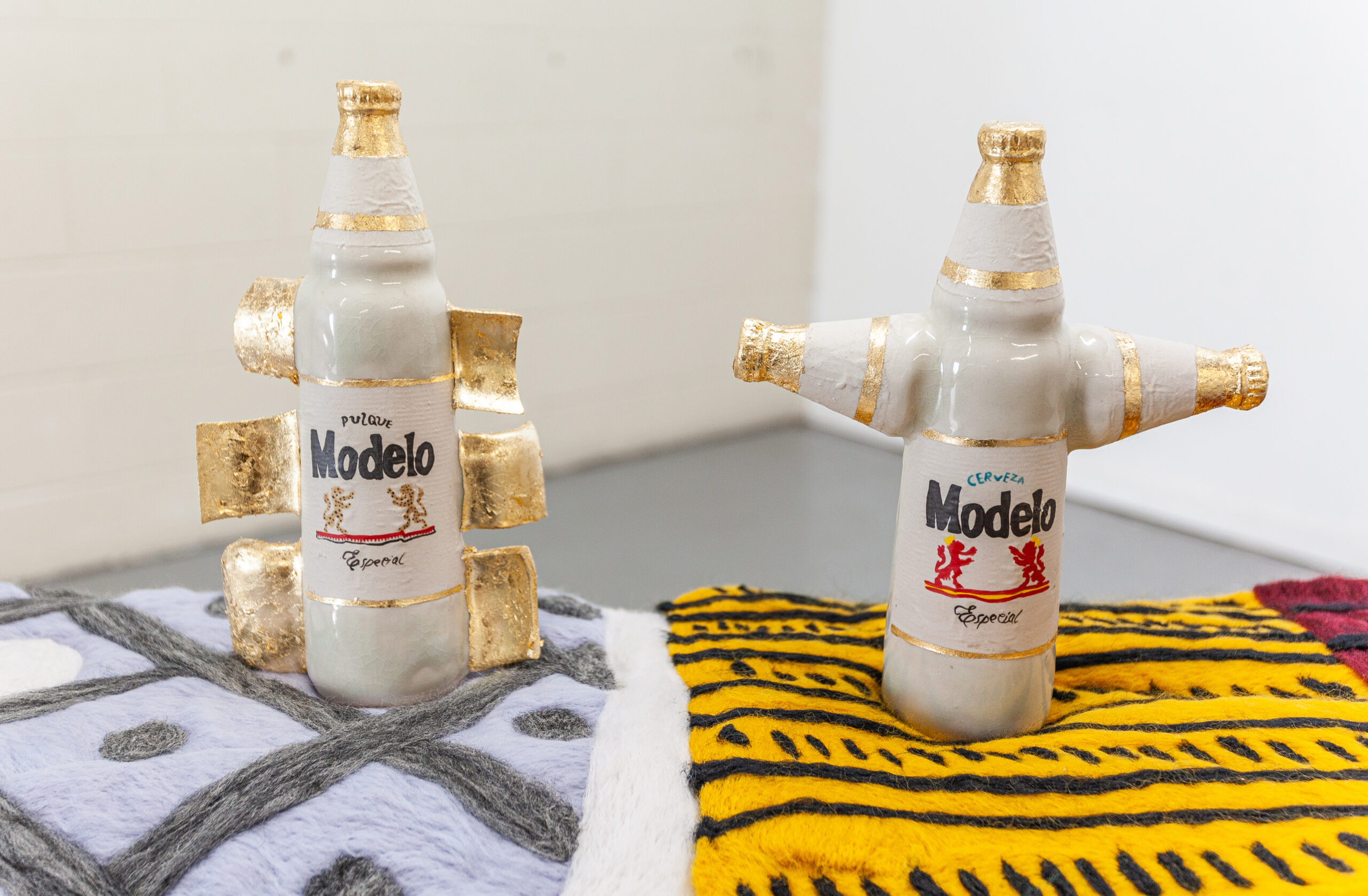 Ceramic Modelo bottles fashioned like a Macuahuitl and cross on felted mats atop a painted pedestal reminiscent of the one that the Mexica gods of life and death stand on.