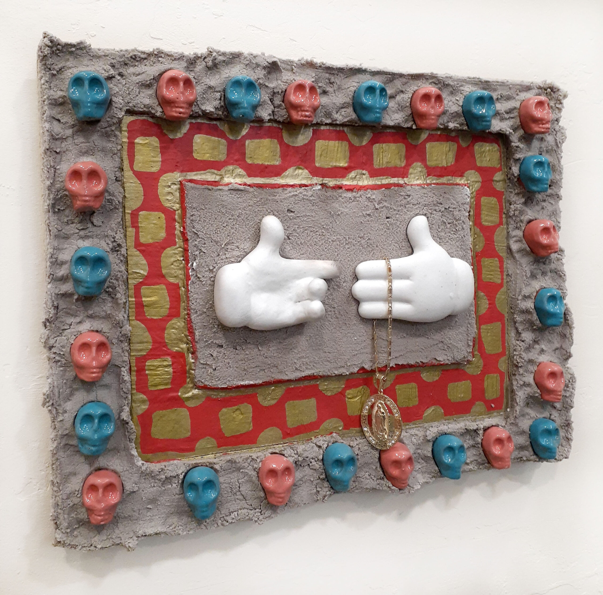 A cement and ceramics relief sculpture featuring the gloves of Mickey Mouse, Mexica skulls, and a gold Virgin Mary necklace.