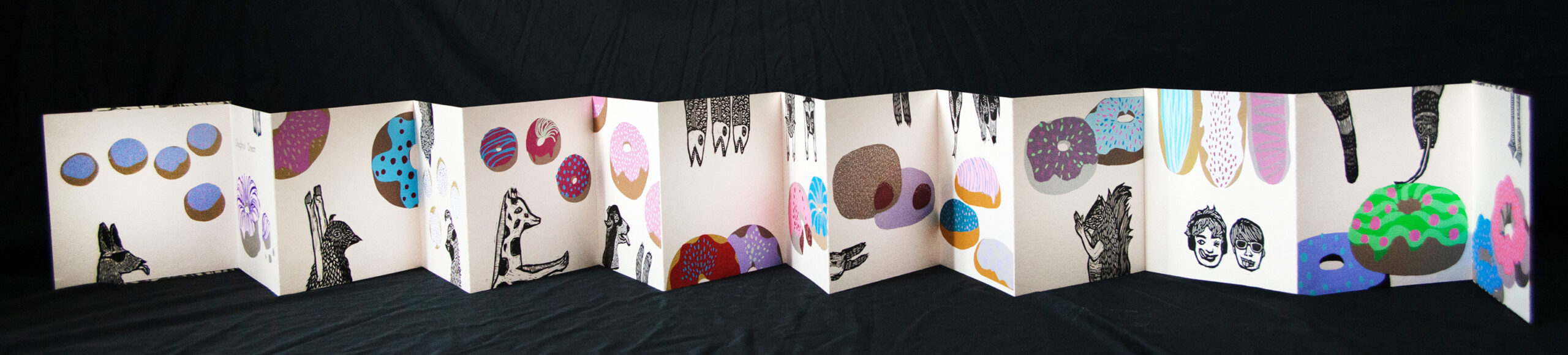 An accordion bound artist book featuring colorful relief printed doughnuts and black and white relief printed humans and animals, each salivating over or reaching for the doughnuts.