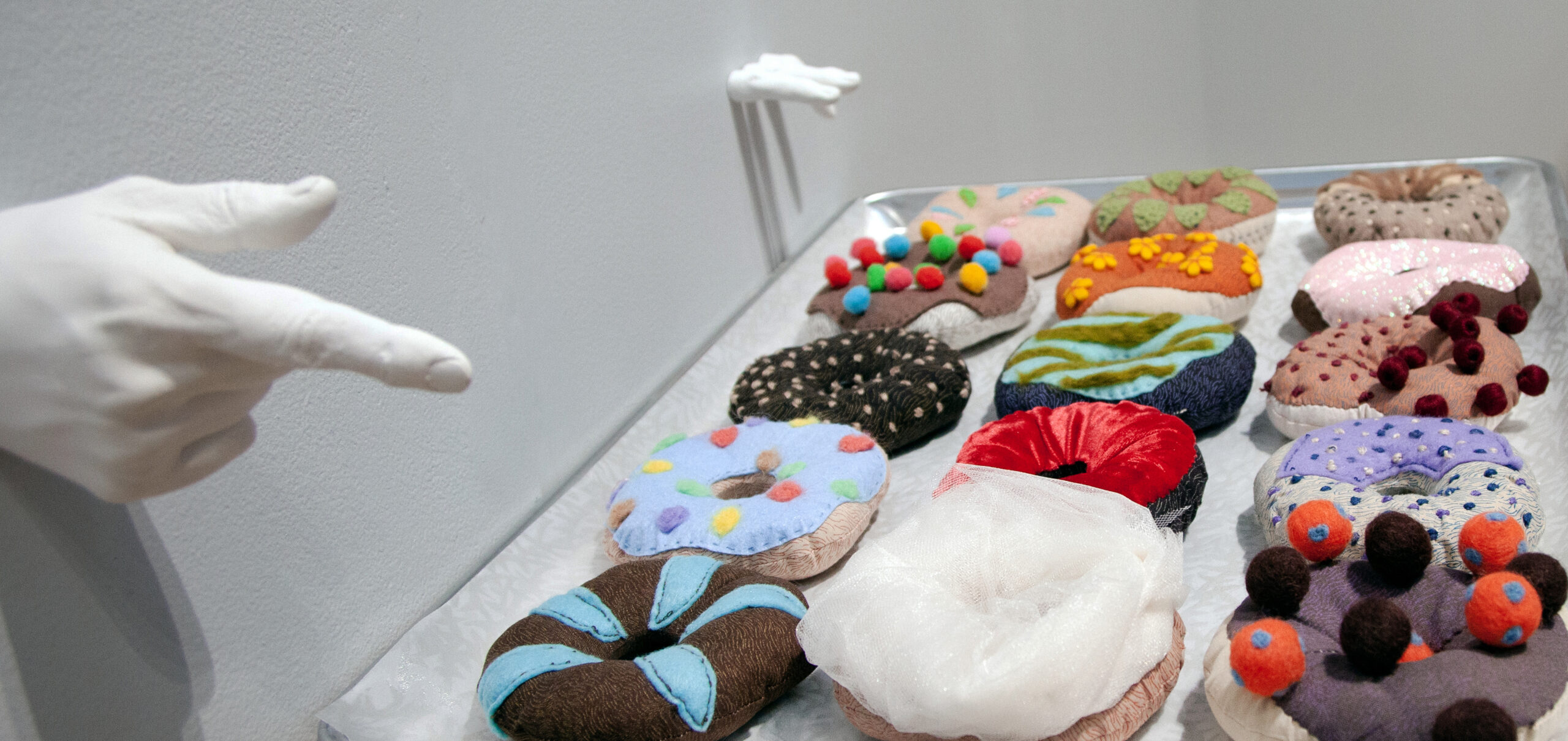 A tray of soft-sculpture doughnuts made from various fabrics, pompoms and accessories. Some fabrics are screen printed with patterned designs. The tray is attached to a gallery wall beneath white plaster casts of fingers and hands in obscene gestures.
