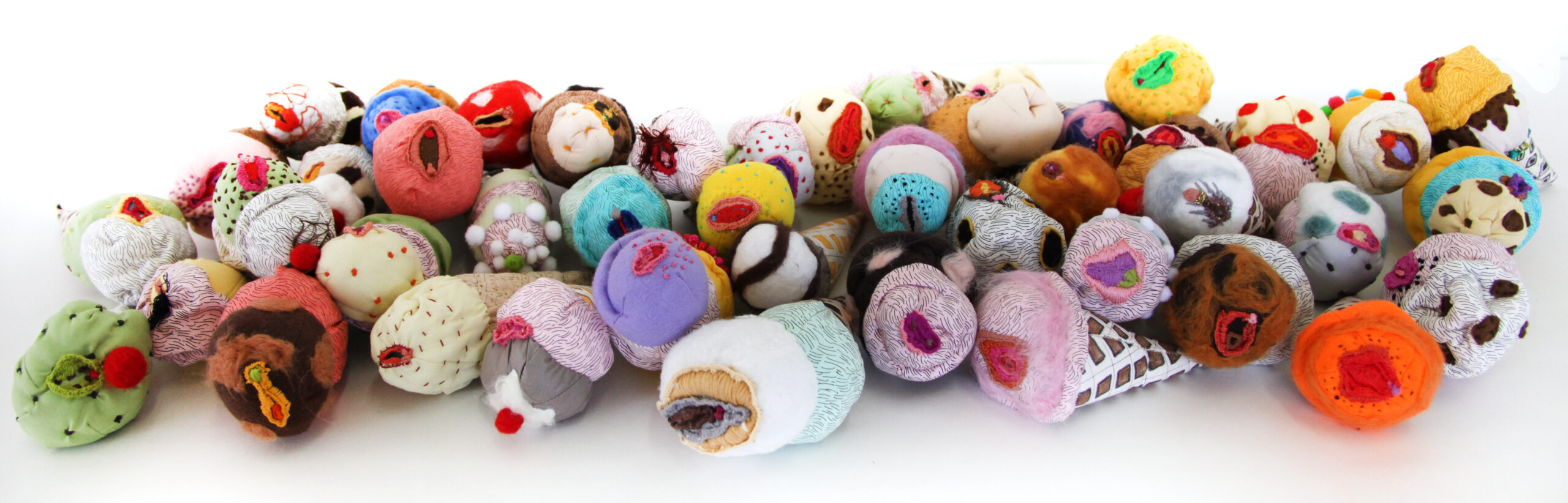 A colorful pile of several dozen soft-sculpture ice cream cones alluding to vaginas and made of various fabrics, pompoms, beads and accessories. Some of the fabrics are screen printed with patterns.