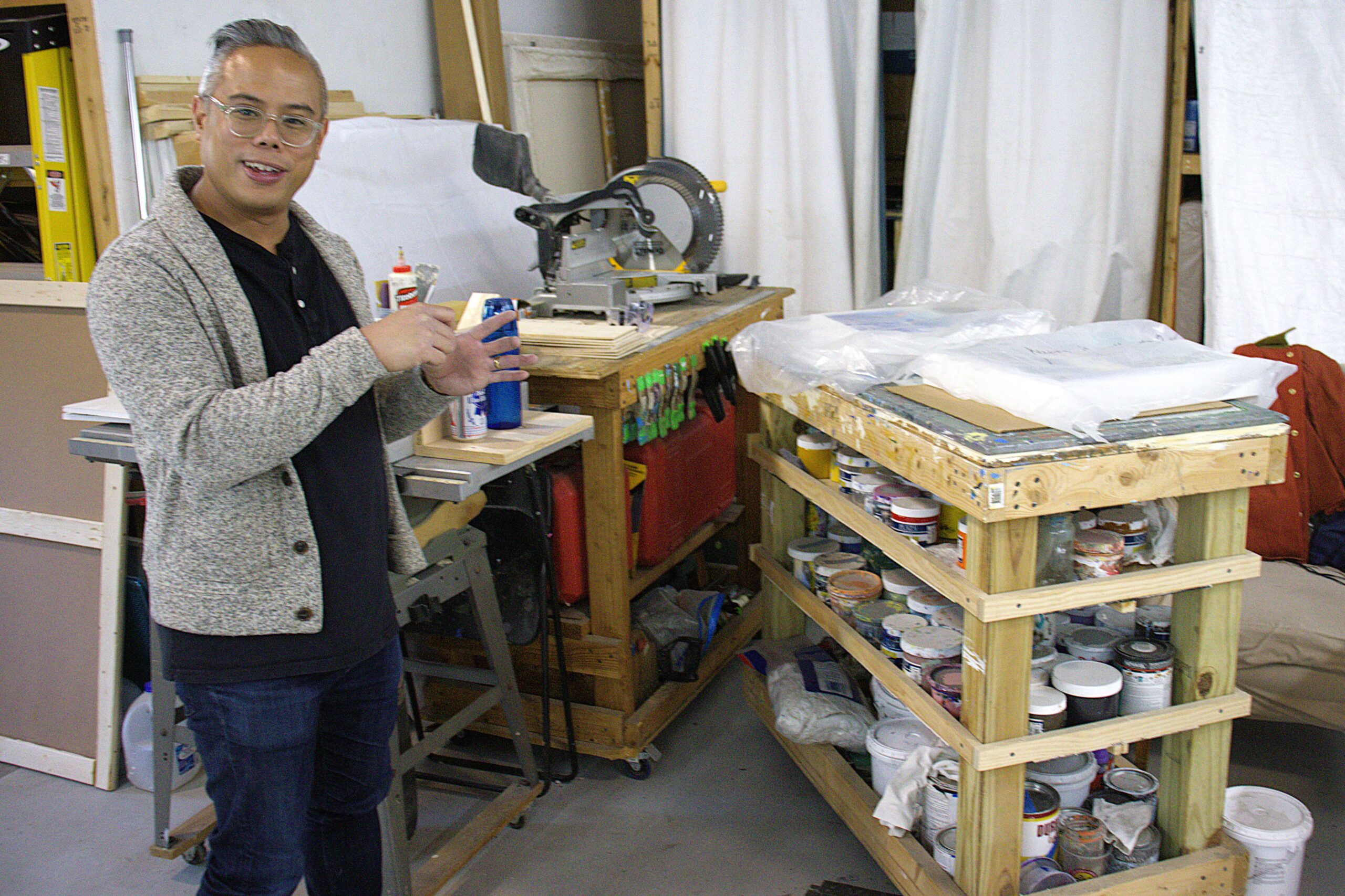 Roberto Jamora stands beside his tools and a shelving unit loaded with various paint containers and pumice medium.