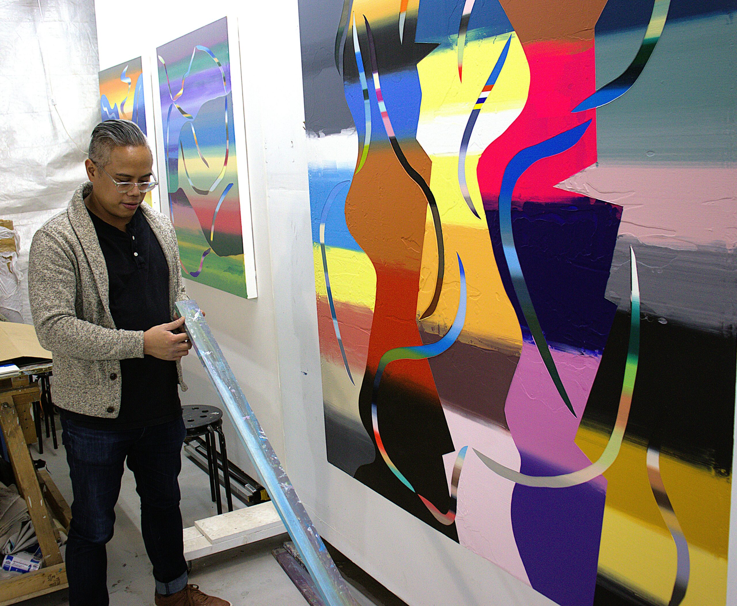 Roberto Jamora holds a large makeshift squeegee made from a length of plywood. He stsands before three of his paintings in his studio space.