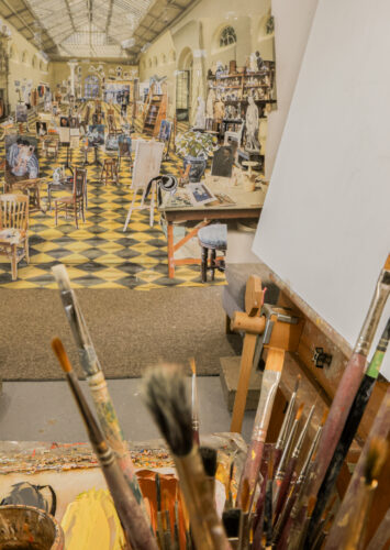 Collage with cut-out paintings and chairs affixed to interior space, with a variety of paintbrushes in front of it