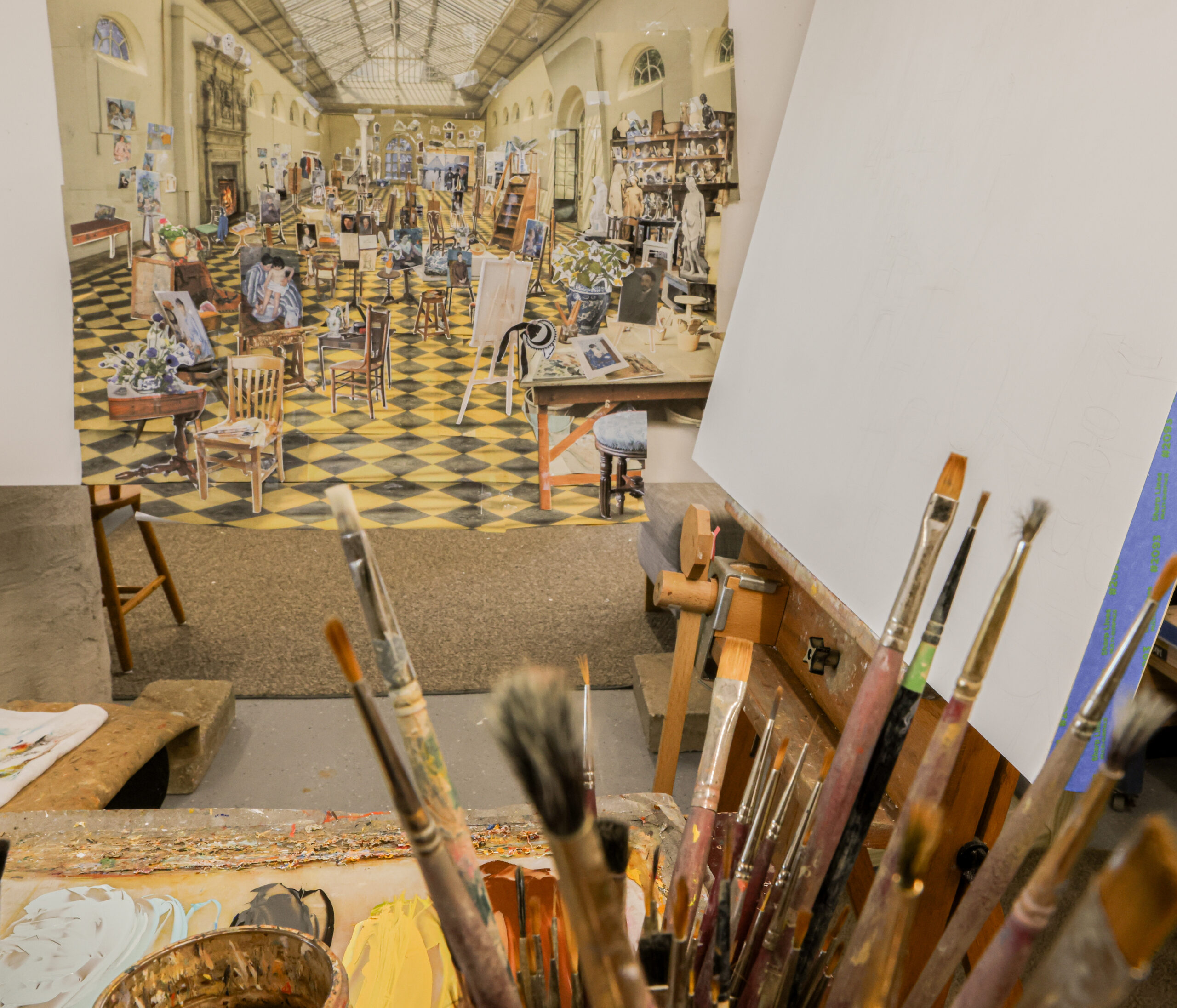 Collage with cut-out paintings and chairs affixed to interior space, with a variety of paintbrushes in front of it