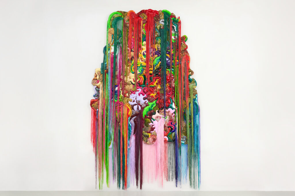 Fabrics in bold colors, including reds and greens, come together in one textile painting with multi-colored fringe hanging both from top and bottom of work.