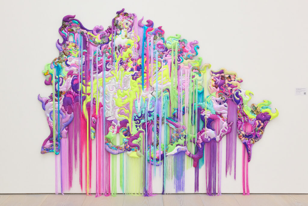 Textiles in a neon color palette (including greens, purple, and pinks) come together to form a woman. Fabrics include florals, sequins, and rainbow patterns. Fringe in various colors hangs down from both higher up the textile painting and the bottom of the textile art.