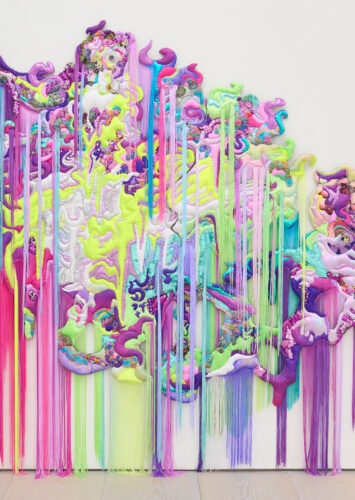 Textiles in a neon color palette (including greens, purple, and pinks) come together to form a woman. Fabrics include florals, sequins, and rainbow patterns. Fringe in various colors hangs down from both higher up the textile painting and the bottom of the textile art.