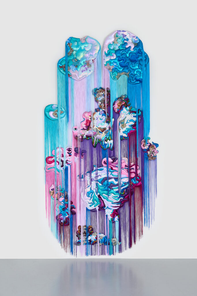 Couple in purple and blue textile collage with mostly blue fabric shapes above them, from which hang blue and light purple fringe. Fringe also hangs from the main portion of textile art.