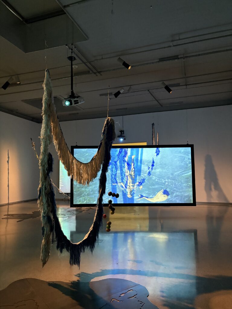 In the foreground, sculpture hanging from ceiling, and in the background, video screen with upside down statue underwater