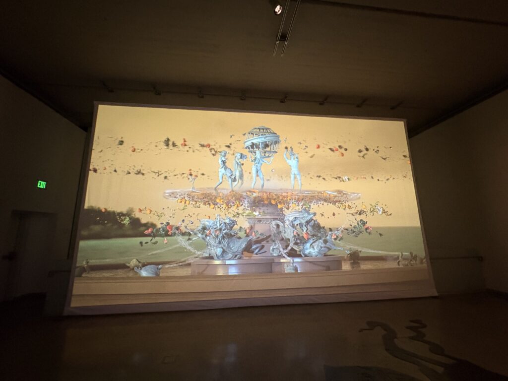 Screen the width of the wall, on which is an image of statues dancing on top of fountain with orange sky in the background