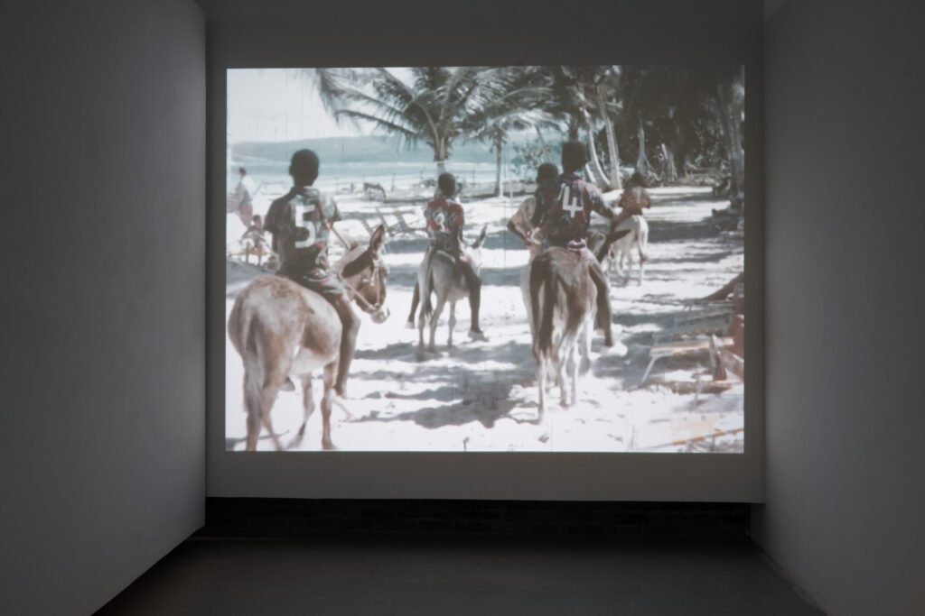 Projected onto the wall, image of boys with numbers on the back of their shirts riding donkeys on the beach
