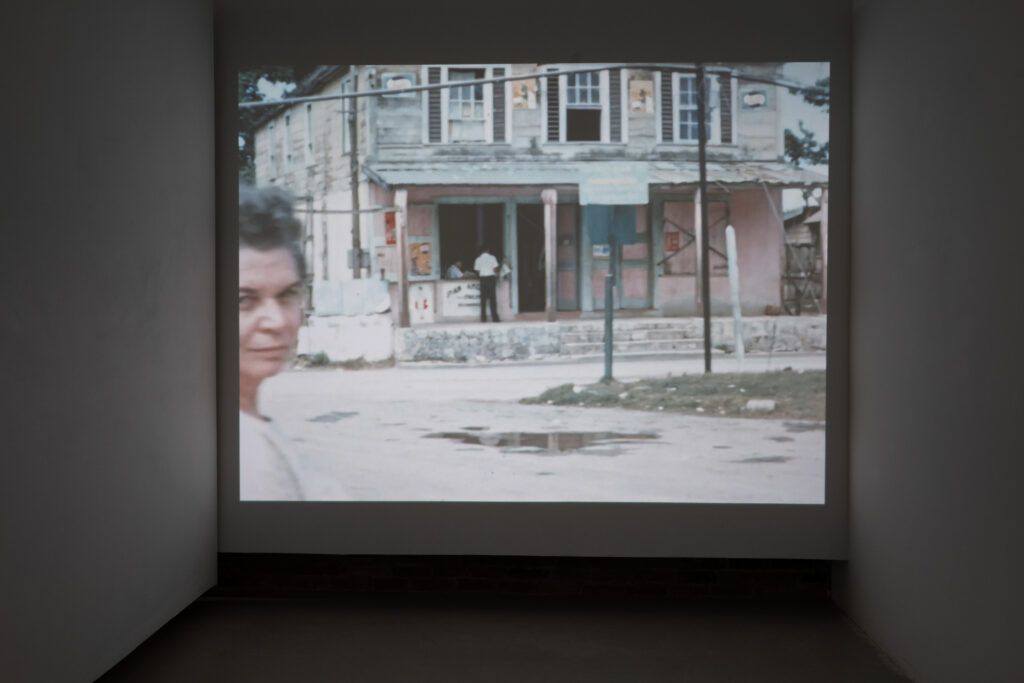 Projected on the wall, woman gazing out at viewer in front of a road and building that has a counter and window with a man sitting behind it and another man facing it.