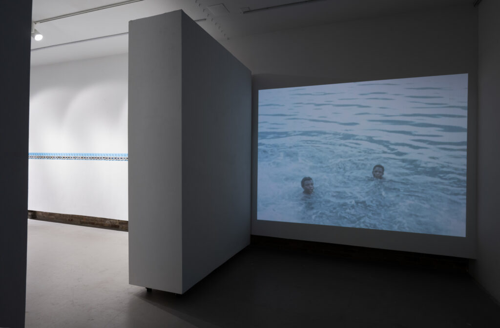 View of gallery showing video projected on one wall and on the other side the Souvenir of Access artwork, a row of keychains on blue. The video still projected on the wall shows two boys or young men's heads sticking out of what looks like ocean water.