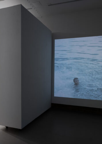 View of gallery showing video projected on one wall and on the other side the Souvenir of Access artwork, a row of keychains on blue. The video still projected on the wall shows two boys or young men's heads sticking out of what looks like ocean water.