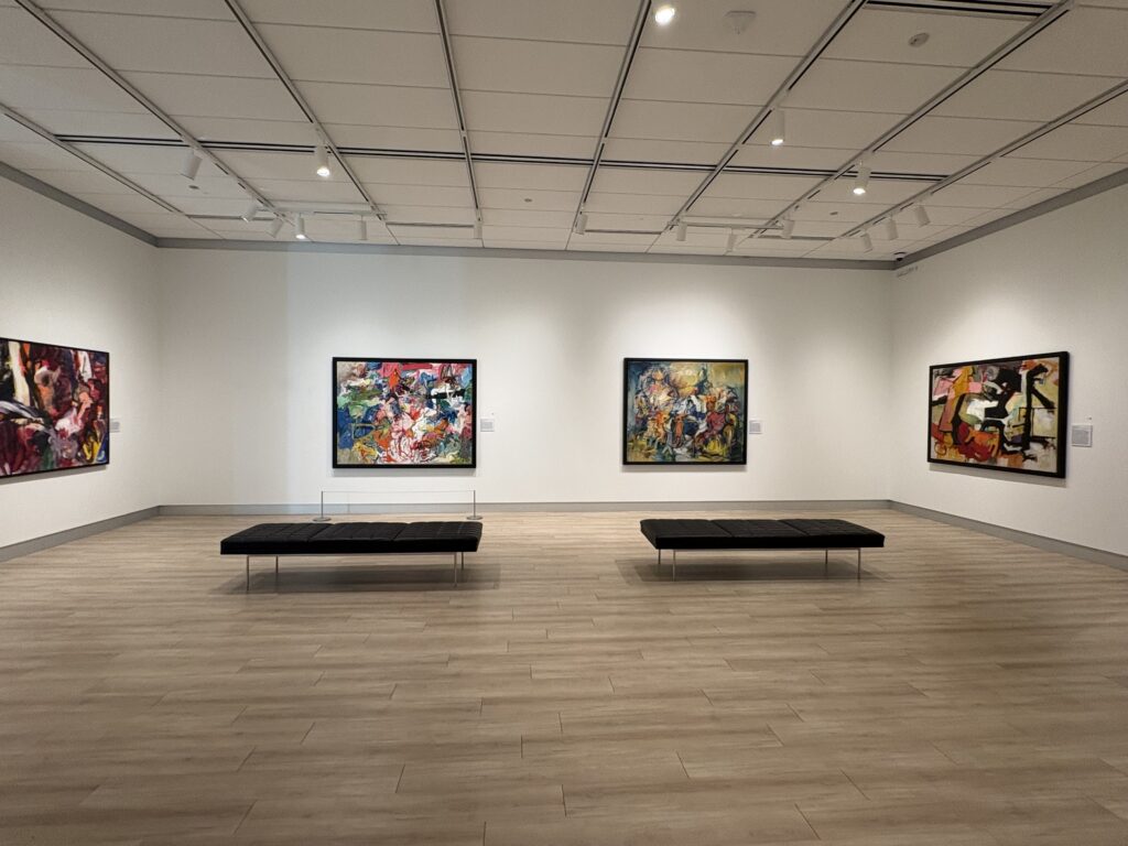 Art gallery with two abstract images on center wall and one each on adjacent walls. Room is well-lit and contains two padded benches to view art.