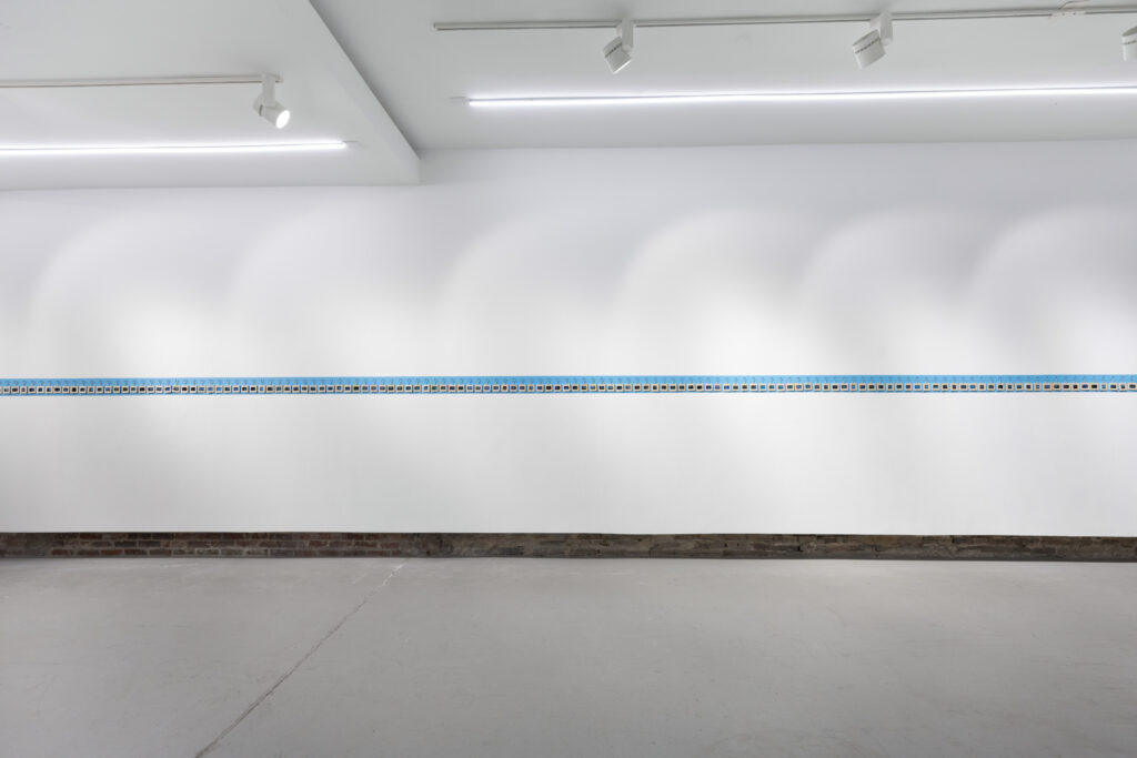 View of whole wall, which shows a blue bar painted horizontally across the white wall. On the bar is a row of hanging square keychains.