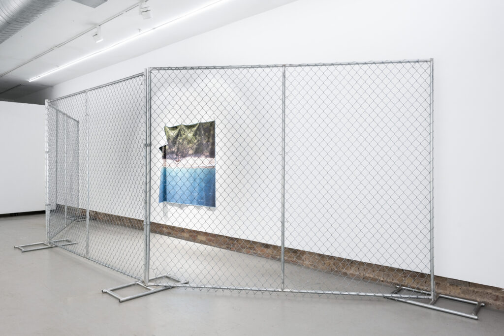 From the side, a chain linked fence, fencing in an image of a beach landscape hanging from a white wall.