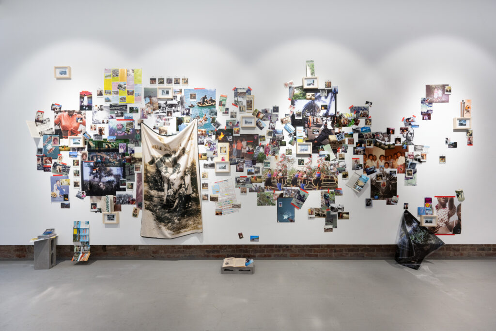 Collage installed across much of a white wall, with photographs, some of which are stuck up by red sticky tape, images printed onto fabric, and props, like a cinder block, a brochure folding out onto the floor, and a book on the floor
