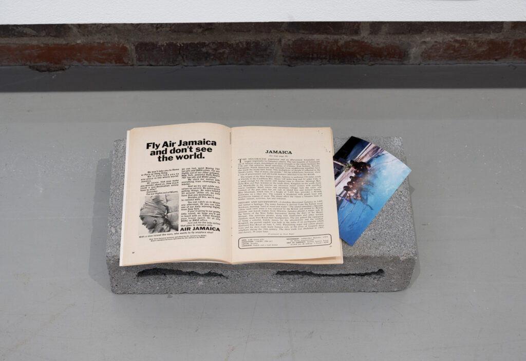 On the floor, a cinder block with a book or catalog on top of it with a title that says "Fly Air Jamaica and don't see the world." The advertisement begins: "We won't take you to Rome or Paris or Hong Kong. We'll only take you one place in the whole world: Jamaica" and ends, "With a nice island like ours: who wants to fly anyplace else?" On the page across from it, there is an article with information about Jamaca, including about its history and government, area, population size, and capital. Next to the book is a personal photo of people in a pool, tilted at an angle.