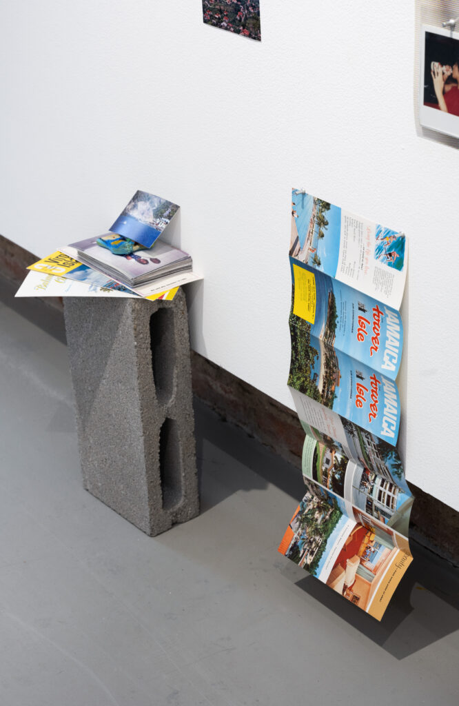 Close-up of installation featuring a brochure unfolded and hanging from its side, almost touching floor, advertising "Jamaica Tower Isle air-conditioned." On a cinder block positioned vertically, a stack of photos and brochures.