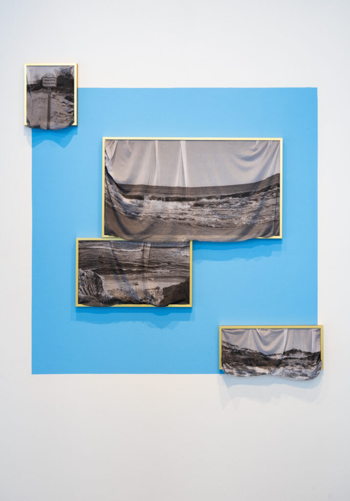 Against blue background, though sometimes extending past blue background, framed images of beach and water. Images are printed onto fabric and are spilling out of gold frames.