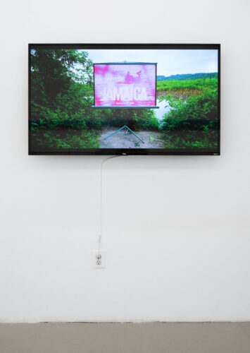 Screen mounted on white wall, showing a still of a video. The image is of a landscape with green plants and water, and within the landscape there is a projector showing an image, pink-hued, of people in a slight, wooden boat with the words "JAMAICA Jamaica Tourist Board" over the image.