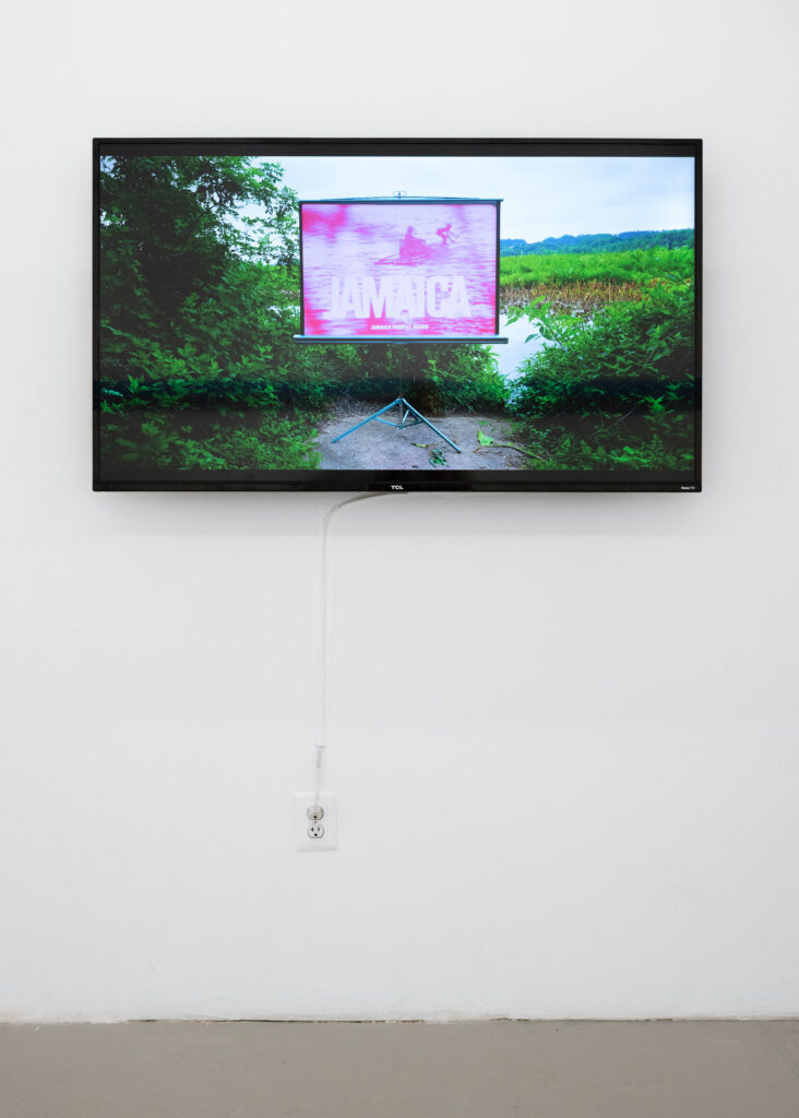 Screen mounted on white wall, showing a still of a video. The image is of a landscape with green plants and water, and within the landscape there is a projector showing an image, pink-hued, of people in a slight, wooden boat with the words "JAMAICA Jamaica Tourist Board" over the image.